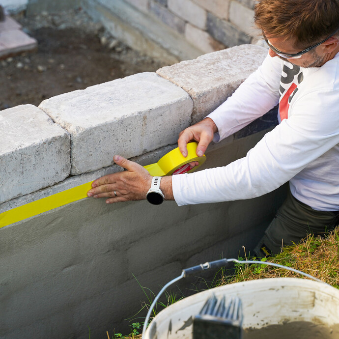 Schütze die umliegenden Bereiche der Gartenmauer erneut mit Klebeband, bevor du die Dichtschlämme aufträgst.